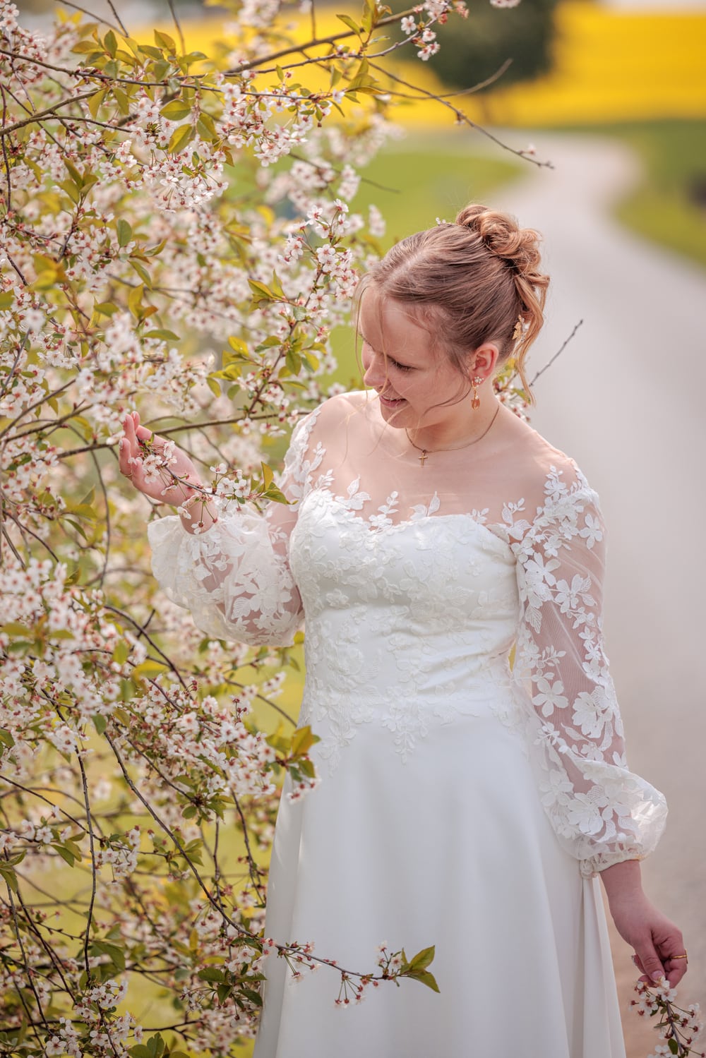 Konfirmand i hvid blondekjole rører ved blomstrende grene med hvide forårsblomster og gul rapsmark i baggrunden. Naturlig konfirmationsfotografering i Randers og Jylland.