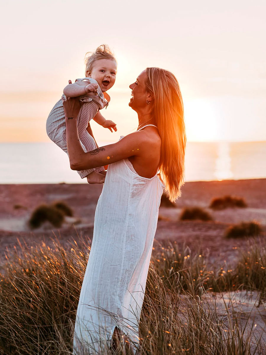 Storslået udendørs familiefotografering af mor og søn i blæsevejret ved Vesterhavet i Jylland. Ægte, livsbekræftende bånd frosset i tid.